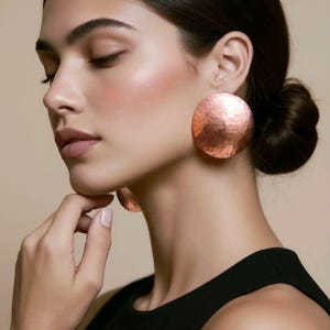 Pode incluir: Grandes brincos circulares de cobre com uma textura martelada distinta. Estas peças de destaque têm um acabamento metálico polido e uma forma arredondada de disco que se assenta no lóbulo, refletindo a luz em toda a sua superfície.