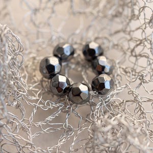 Pu&ograve; includere: Una disposizione circolare di sei perline metalliche grigio scuro sfaccettate, poggiate all'interno di una delicata struttura a rete di filo color argento. Le perline presentano una superficie lucida e riflettente disposte ad anello.