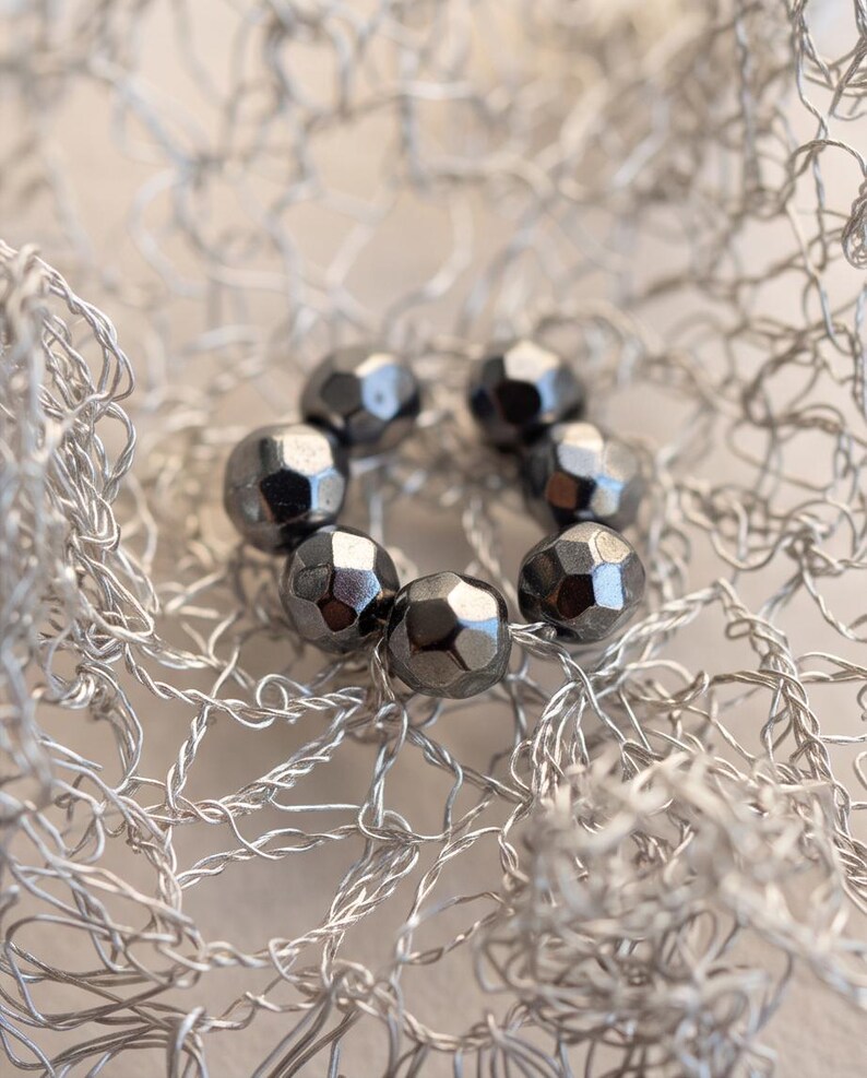 Pu&ograve; includere: Una disposizione circolare di sei perline metalliche grigio scuro sfaccettate, poggiate all'interno di una delicata struttura a rete di filo color argento. Le perline presentano una superficie lucida e riflettente disposte ad anello.