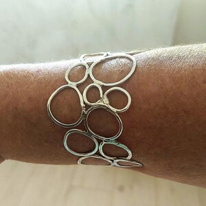 May include: A silver-toned cuff bracelet featuring an openwork design of interconnected, irregular oval shapes. The bracelet is worn on a wrist, showcasing its modern, artistic style. The metal has a polished finish, reflecting light.