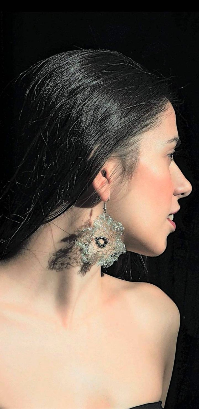 Pu&ograve; includere: Un orecchino pendente caratterizzato da una grande e complessa forma floreale simile al pizzo, realizzata in una sottile rete metallica color argento scintillante. Il centro del fiore &egrave; accentuato da una piccola perla circolare scura.
