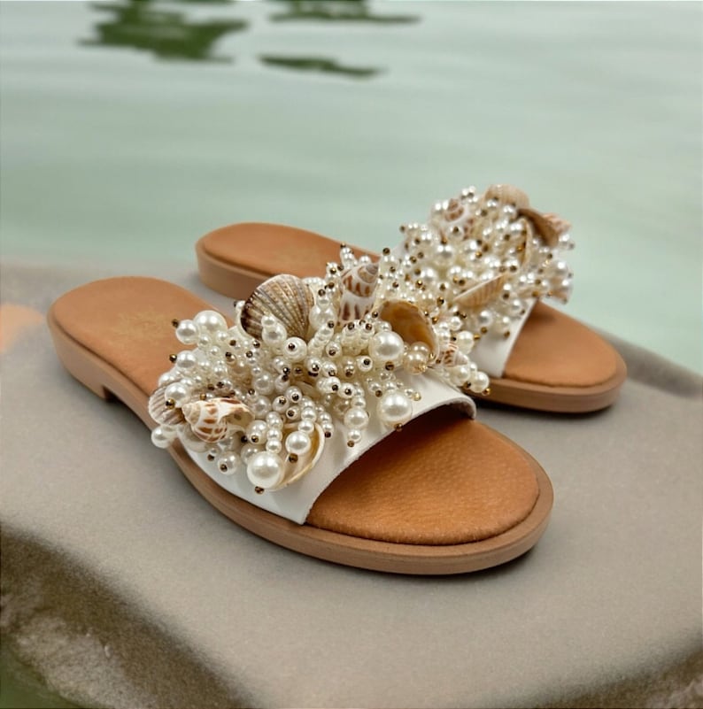 May include: White leather sandals with a large embellishment of faux pearls and seashells. The sandals have a brown leather footbed.