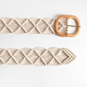 Puede incluir: Cinturón de macramé color crema con un diseño tejido y una hebilla redonda de tono natural. El intrincado patrón del cinturón presenta formas de diamante y líneas curvas, creando una apariencia texturizada. La hebilla tiene un pasador de metal dorado.