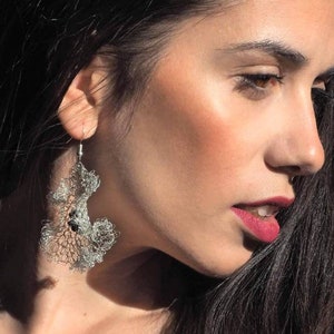 Pu&ograve; includere: Un orecchino pendente color argento caratterizzato da un intricato design a rete metallica traforata che ricorda strutture organiche di corallo o pizzo. L'orecchino &egrave; sospeso a un semplice gancio in metallo e presenta piccoli accenti circolari scuri.