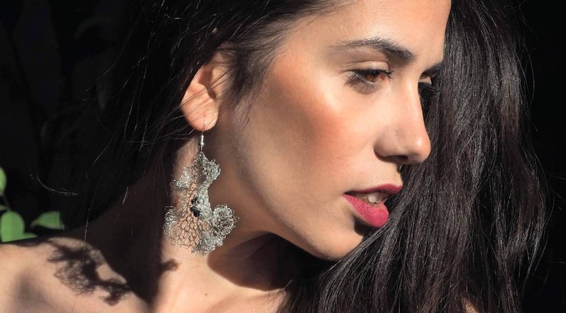 Pu&ograve; includere: Un orecchino pendente color argento caratterizzato da un intricato design a rete metallica traforata che ricorda strutture organiche di corallo o pizzo. L'orecchino &egrave; sospeso a un semplice gancio in metallo e presenta piccoli accenti circolari scuri.