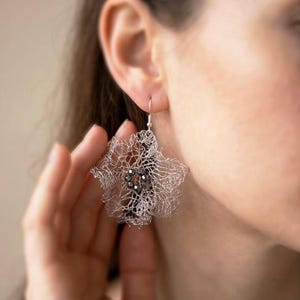 Pu&ograve; includere: Un orecchino in rete metallica color argento a forma di fiore delicato e arricciato. Il centro dell'orecchino presenta un gruppo di piccole perline scure sfaccettate. L'orecchino pende da un semplice gancio color argento.