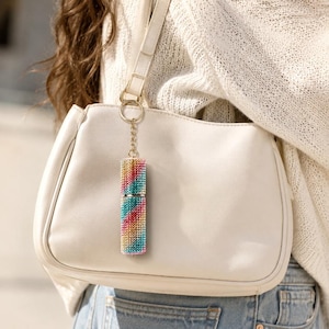 Puede incluir: Un dije atomizador de perfume cilíndrico cubierto con rayas diagonales de pedrería multicolor, incluyendo rosa, amarillo y azul, unido a una cadena y anillo de metal dorado, colgando de la correa de un bolso de hombro liso color blanco hueso.