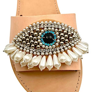 May include: A light pink leather sandal with an eye-shaped embellishment. The eye design features a black center, surrounded by blue and clear crystals, silver beads, and white teardrop-shaped pearls. The sandal has a simple strap design.