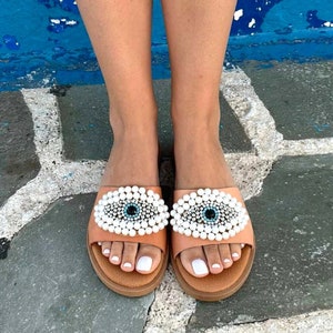 May include: A pair of tan leather sandals with white pearl embellishments and a blue evil eye design. The sandals have a simple, slip-on style.