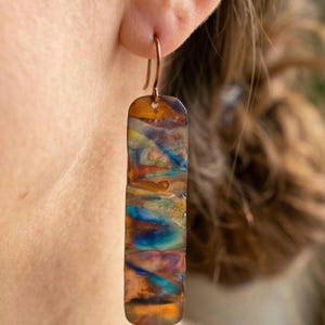 Puede incluir: Un pendiente rectangular colgante con una superficie de metal tratada térmicamente que presenta patrones en espiral de colores azul, naranja, amarillo y marrón. El pendiente cuelga de un gancho cobrizo y tiene una forma alargada y plana.