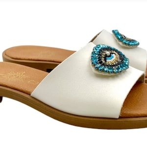 Op de afbeelding: Witte leren instappers met bruine zolen en voetbedden. Elke sandaal heeft een decoratief blauw en goud kralen oogontwerp op de band. Deze sandalen zijn geschikt voor casual wear.