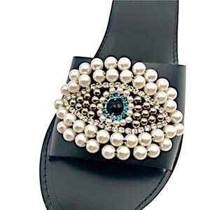 May include: Black leather slide sandals with a pearl and rhinestone embellished evil eye design.