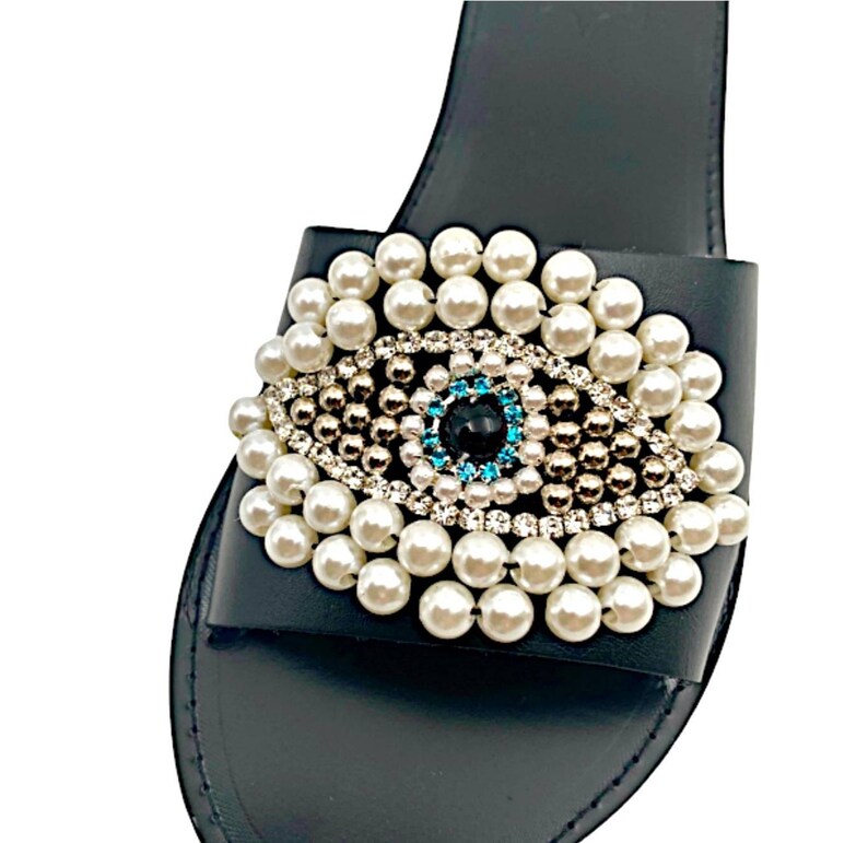 May include: Black leather slide sandals with a pearl and rhinestone embellished evil eye design.