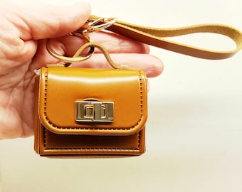 May include: A small, mustard-yellow, rectangular pouch with a smooth finish and a gold-tone metal turn-lock closure on the front flap. It features a matching mustard-yellow wrist strap attached to a gold-tone metal split ring key holder.