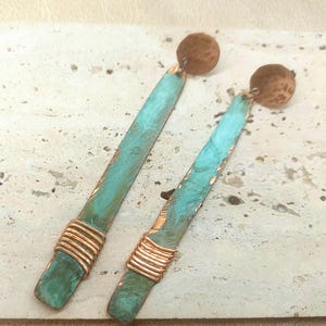 Puede incluir: Un par de pendientes largos, finos y rectangulares con un acabado de pátina de color turquesa sobre metal. Cada pendiente tiene una parte superior circular de cobre martillado y una sección de alambre de cobre envuelta alrededor de la base.