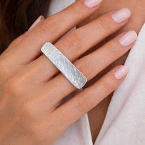 Könnte beinhalten: Ein silberner Ring mit rechteckigem, gehämmertem Design. Der Ring wird an einem Finger getragen und zeigt seine strukturierte Oberfläche und seinen metallischen Glanz. Das Design des Rings ist schlicht, aber elegant und für den Alltag geeignet.