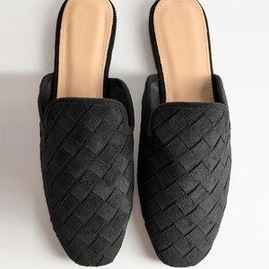 May include: A pair of black woven slip-on mules. The shoes have a textured, basket-weave design on the upper and a light tan insole. The mules are set against a white background.