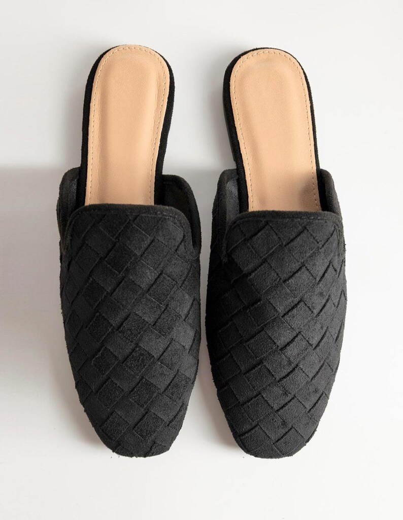 May include: A pair of black woven slip-on mules. The shoes have a textured, basket-weave design on the upper and a light tan insole. The mules are set against a white background.