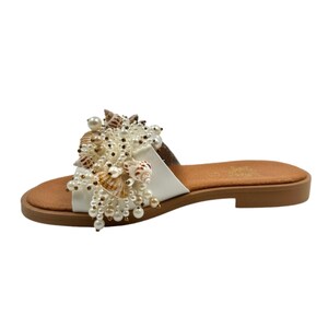 May include: White leather slide sandals with a large embellishment of seashells and pearls.