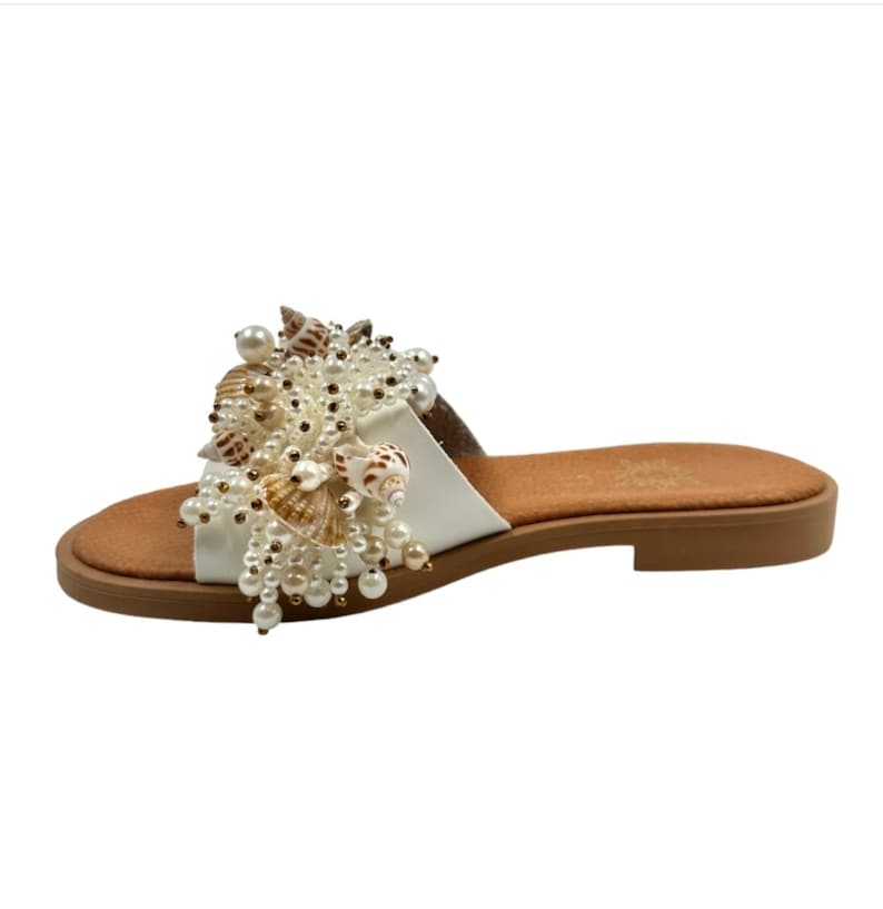 May include: White leather slide sandals with a large embellishment of seashells and pearls.