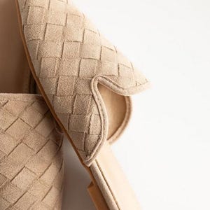 May include: Close-up of beige woven suede slip-on shoes. The shoes have a pointed toe and a low heel. The woven pattern covers the entire upper part of the shoe. The sole is a light brown color. The background is white.