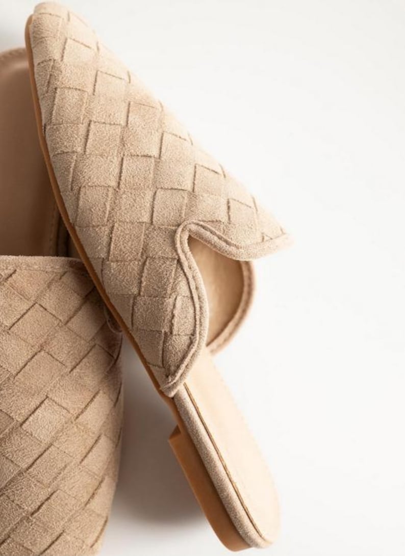 May include: Close-up of beige woven suede slip-on shoes. The shoes have a pointed toe and a low heel. The woven pattern covers the entire upper part of the shoe. The sole is a light brown color. The background is white.