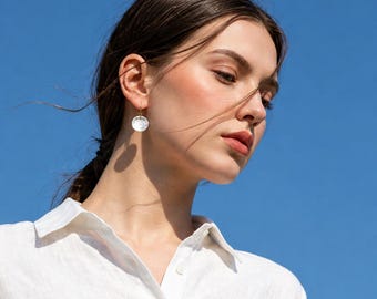 May include: A single circular earring featuring a white, iridescent mother-of-pearl disc suspended from a thin gold-toned metal hook. The earring is worn on the earlobe, showcasing a smooth, reflective surface that catches the natural light.