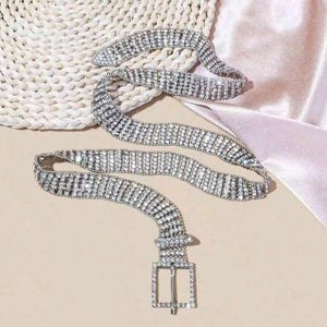 May include: A silver-toned belt covered in multiple rows of clear, sparkling rhinestones. The belt features a matching square-shaped buckle frame, also encrusted with rhinestones, and a prong for fastening. The design consists of several parallel strands.