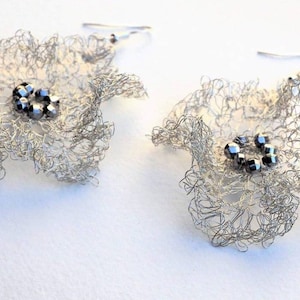Pu&ograve; includere: Un paio di orecchini in filo metallico argentato a forma di fiore. Ogni orecchino presenta una delicata rete traforata di filo d'argento sottile che forma petali arricciati, con un gruppo di piccole perline metalliche scure al centro.