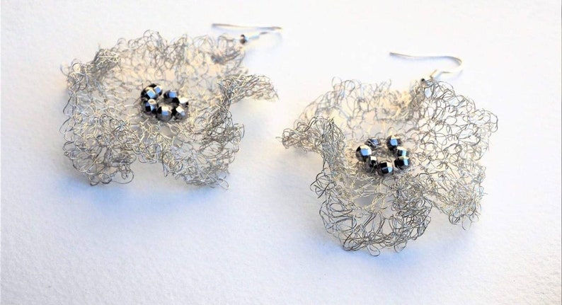 Pu&ograve; includere: Un paio di orecchini in filo metallico argentato a forma di fiore. Ogni orecchino presenta una delicata rete traforata di filo d'argento sottile che forma petali arricciati, con un gruppo di piccole perline metalliche scure al centro.