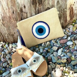 May include: A beige clutch with a large blue and white eye design, paired with white leather sandals. The sandals feature an eye design with decorative embellishments. The items are displayed on a bed of pebbles.
