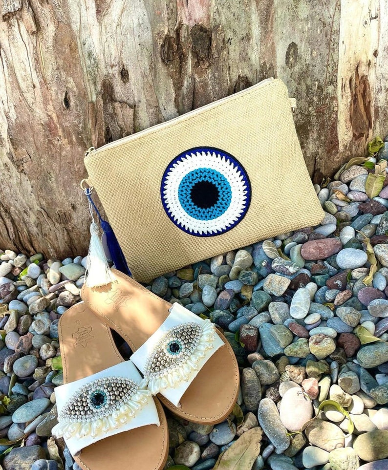 May include: A beige clutch with a large blue and white eye design, paired with white leather sandals. The sandals feature an eye design with decorative embellishments. The items are displayed on a bed of pebbles.