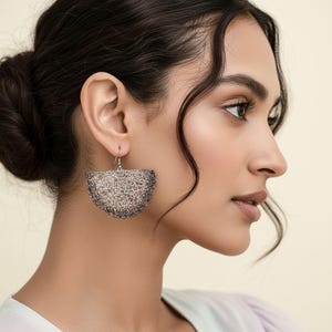 Puede incluir: Primer plano de una mujer con un pendiente de malla de alambre plateado y gris. El pendiente tiene forma de semicírculo con una superficie texturizada y un gancho plateado. La mujer tiene el pelo oscuro y lleva una blusa clara.