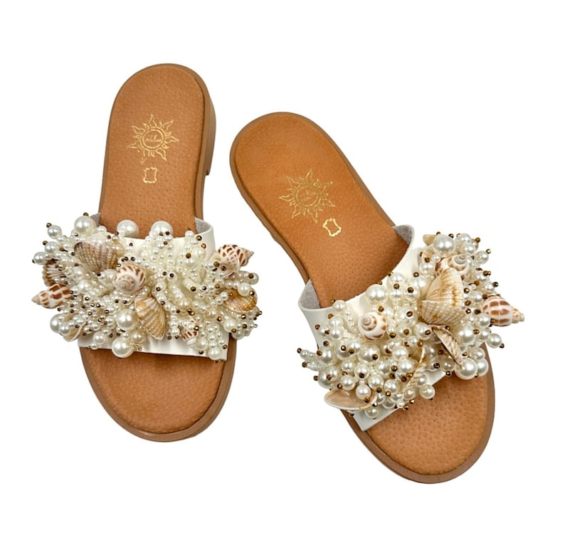 May include: White leather sandals with a brown sole and a large cluster of white pearls and seashells on the front strap.
