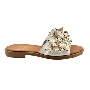 May include: White leather slide sandals with a large, decorative cluster of faux pearls and seashells. The sandals have a brown platform sole.