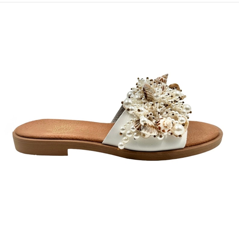 May include: White leather slide sandals with a large, decorative cluster of faux pearls and seashells. The sandals have a brown platform sole.