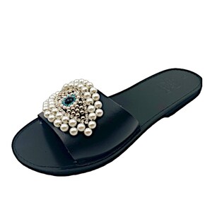 May include: Black leather slide sandals with a pearl and crystal embellished evil eye design.