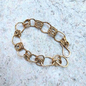 Op de afbeelding: Een goudkleurige armband met een uniek kettingontwerp. De armband heeft een reeks onderling verbonden ovale schakels, afgewisseld met decoratieve, gewikkelde segmenten. De sluiting is een eenvoudige haak- en oogsluiting. De sieraden worden gepresenteerd op een getextureerd, lichtgekleurd oppervlak.