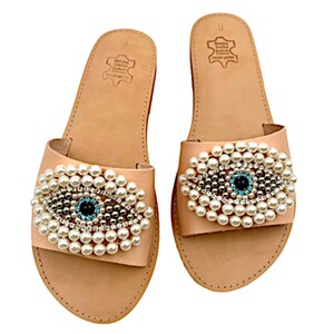 May include: A pair of light brown leather sandals with a pearl and crystal embellished evil eye design. The sandals have a simple, flat design and are perfect for summer.