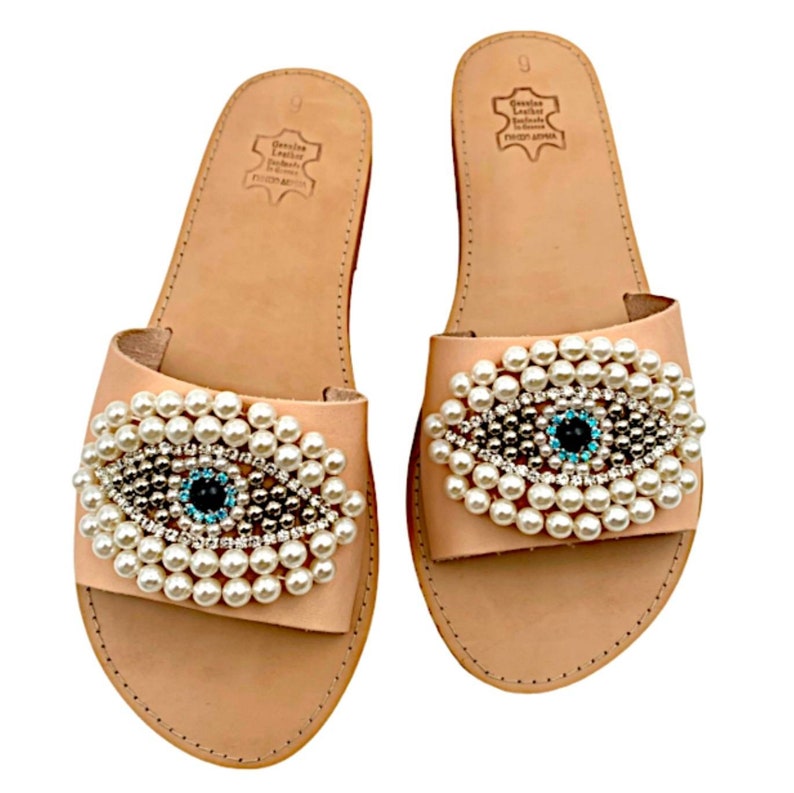 May include: A pair of light brown leather sandals with a pearl and crystal embellished evil eye design. The sandals have a simple, flat design and are perfect for summer.