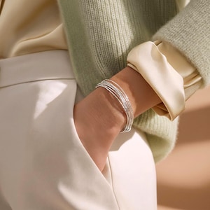 Op de afbeelding: Een zilveren armband met meerdere strengen, bestaande uit verschillende dunne, getextureerde metalen ringen die om de pols vallen. De armband heeft een gepolijste, reflecterende afwerking en wordt gedragen met een lichtgroen gebreid vest en een crèmekleurig satijnen overhemd.
