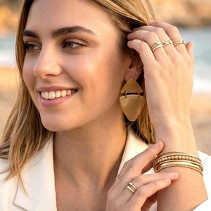 Peut inclure: Un ensemble de bijoux dorés comprenant une paire de boucles d'oreilles pendantes géométriques avec une partie supérieure triangulaire et une partie inférieure arrondie, plusieurs bracelets fins dorés et plusieurs bagues dorées.