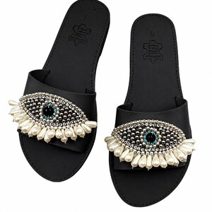 May include: A pair of black leather sandals with an eye-shaped embellishment. The eye design features a black center, surrounded by silver beads and white pearl accents. The sandals are size 9.