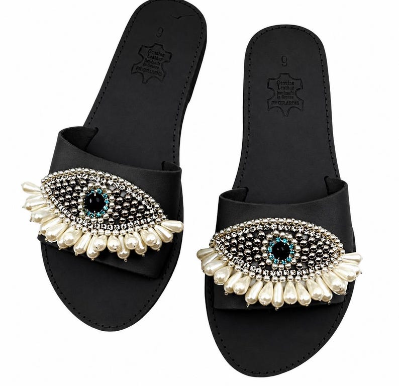 May include: A pair of black leather sandals with an eye-shaped embellishment. The eye design features a black center, surrounded by silver beads and white pearl accents. The sandals are size 9.