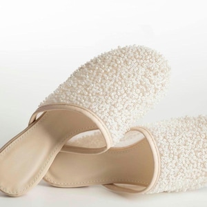 May include: A pair of slip-on mules featuring a rounded toe and a beige inner sole. The upper part of the footwear is covered in dense, small white beads and sequins, creating a textured, shimmering surface that contrasts with the smooth beige trim.