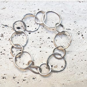 Op de afbeelding: Zilverkleurige armband met een reeks onderling verbonden, gehamerde cirkels. De armband is in een cirkelvormig patroon gerangschikt, waarbij de schakels en het licht dat van het metaal weerkaatst, worden benadrukt. De sieraden worden gepresenteerd op een getextureerd, lichtgekleurd oppervlak.