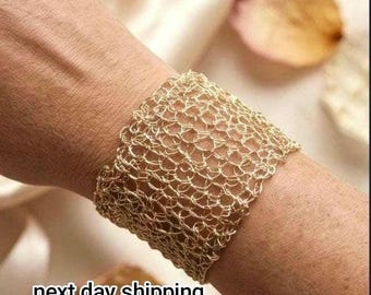 Può includere: Un bracciale a fascia larga realizzato in filo sottile color oro intrecciato in un motivo a rete aperta. Il bracciale presenta una struttura flessibile e complessa che avvolge il polso, mettendo in mostra un design metallico delicato.