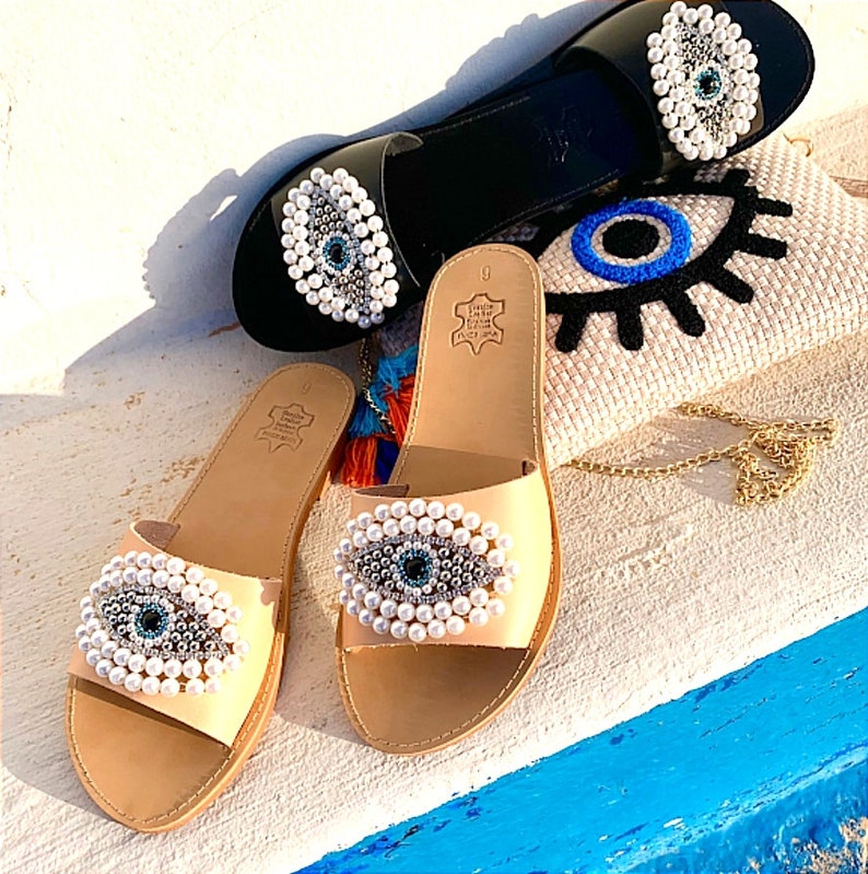 May include: Two pairs of sandals with a blue evil eye design. The sandals are made of leather and have a beaded evil eye design. The sandals are on a white surface.