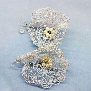 Pu&ograve; includere: Un paio di orecchini in rete metallica color argento a forma di fiori aperti. Ogni orecchino presenta una delicata e complessa trama di sottile filo metallico che forma una struttura a petali, con un gruppo di piccole perle color crema al centro.