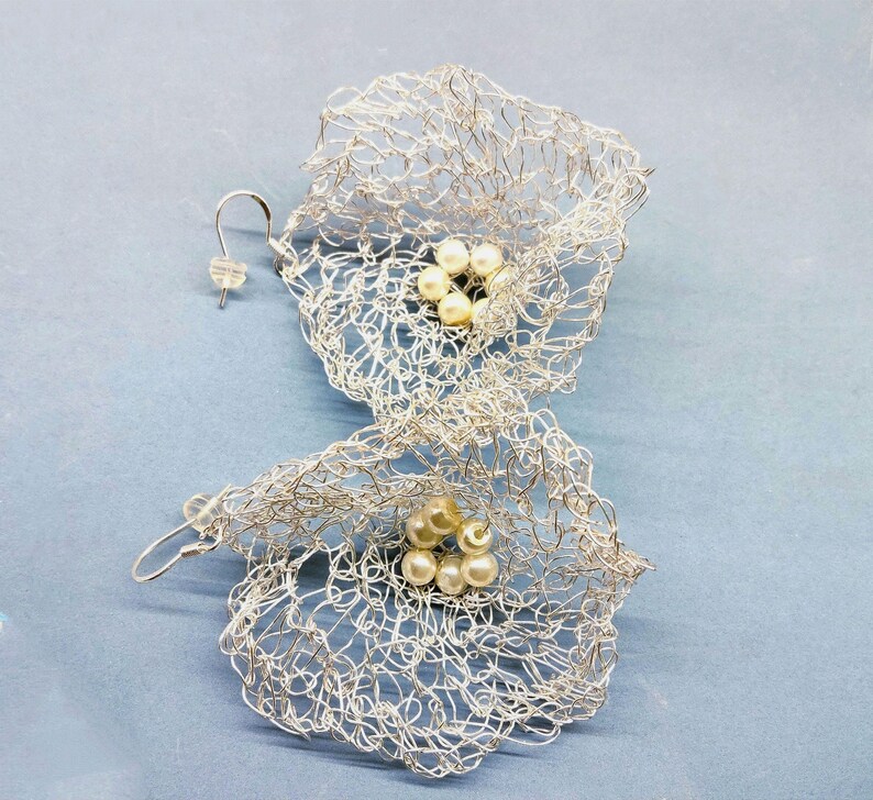Pu&ograve; includere: Un paio di orecchini in rete metallica color argento a forma di fiori aperti. Ogni orecchino presenta una delicata e complessa trama di sottile filo metallico che forma una struttura a petali, con un gruppo di piccole perle color crema al centro.